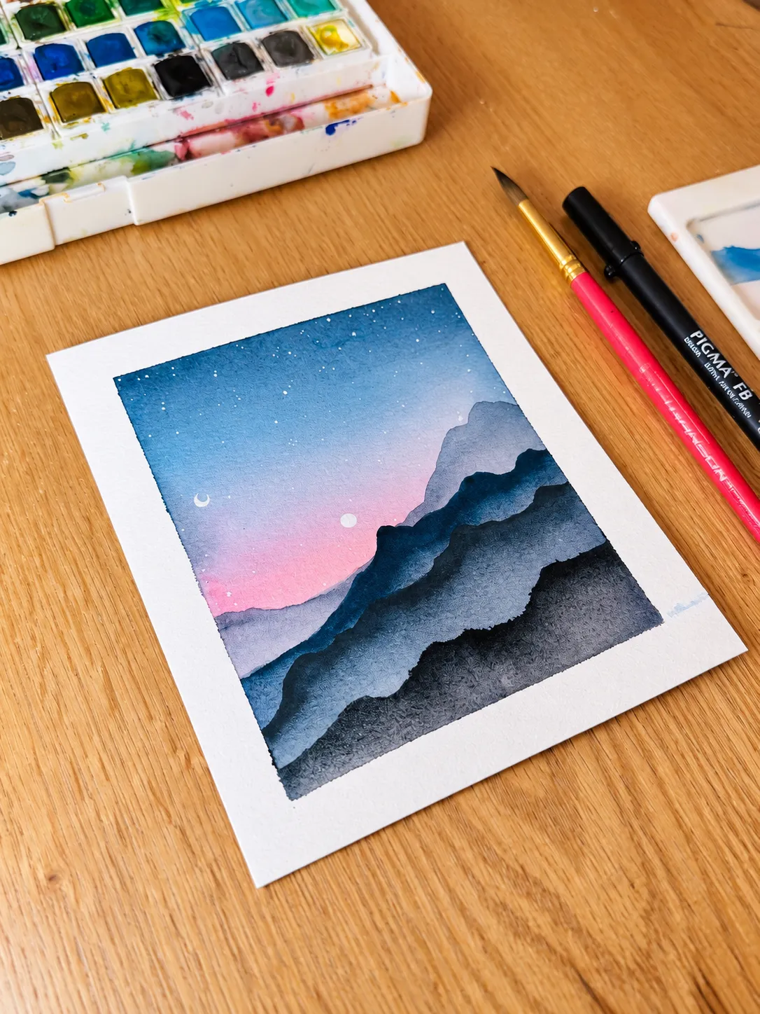 Blue mountain watercolor beside a palette and brushes