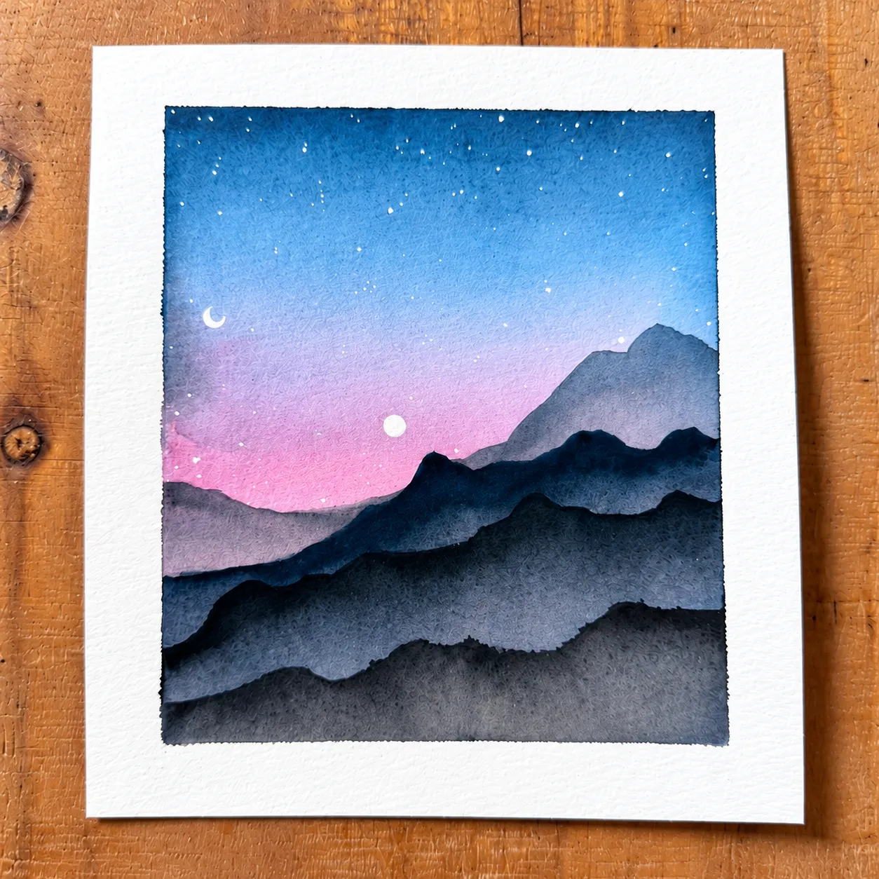 Blue mountain watercolor beneath a starry sky lying on a wooden table