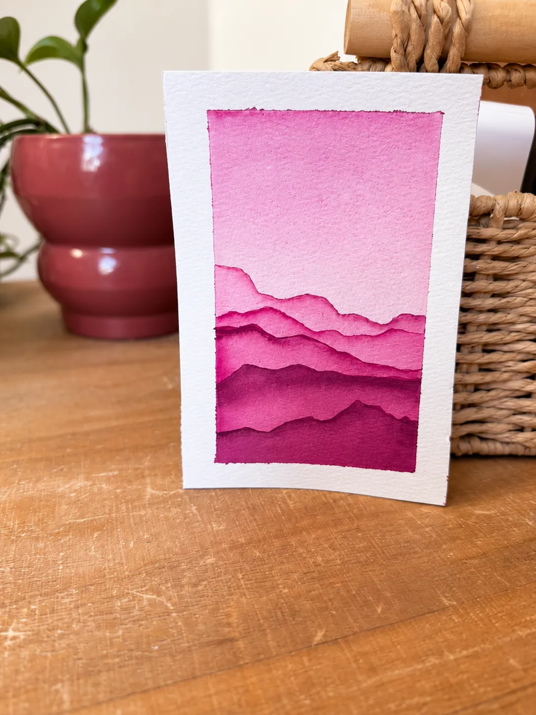 Layered pink mountain watercolor standing beside a plant and woven basket